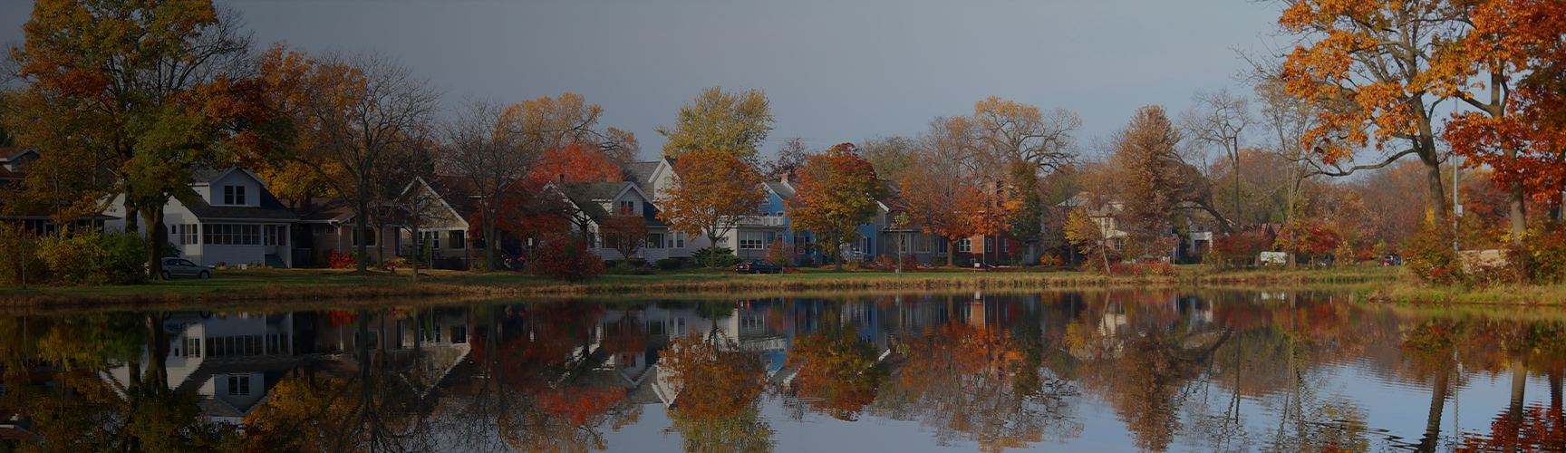 Neighborhood homes and autumnal trees are mirrored in a calm pond.